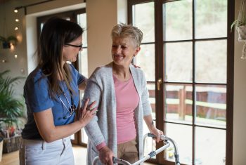 Healthcare worker or caregiver visiting senior woman indoors at home, helping her to walk Healthcare worker or caregiver visiting senior woman indoors at home, helping her to walk