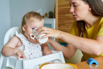 Mother feeding daughter with baby formula in mug. Healthy food. Child care Mother feeding daughter with baby formula in mug. Healthy food. Child care